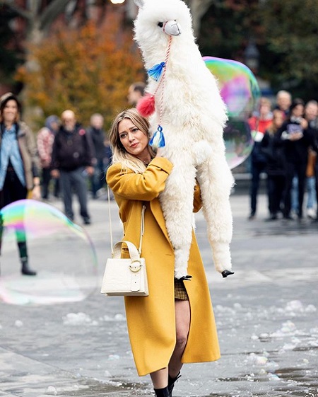 Hilary Duff in a trench coat holding a white stuffed alpaca.